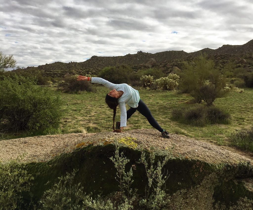 Blythe doing yoga Wild Thing pose of a rock in the desert on a cloudy day with mountains and trees and cactus in the background