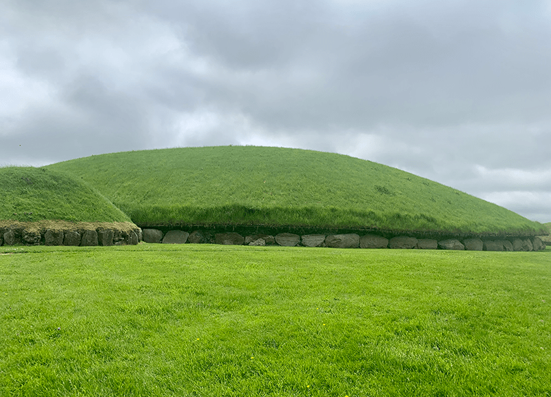 Two bright green sidhe mounds from the Knowth complex against a grey, cloudy sky.