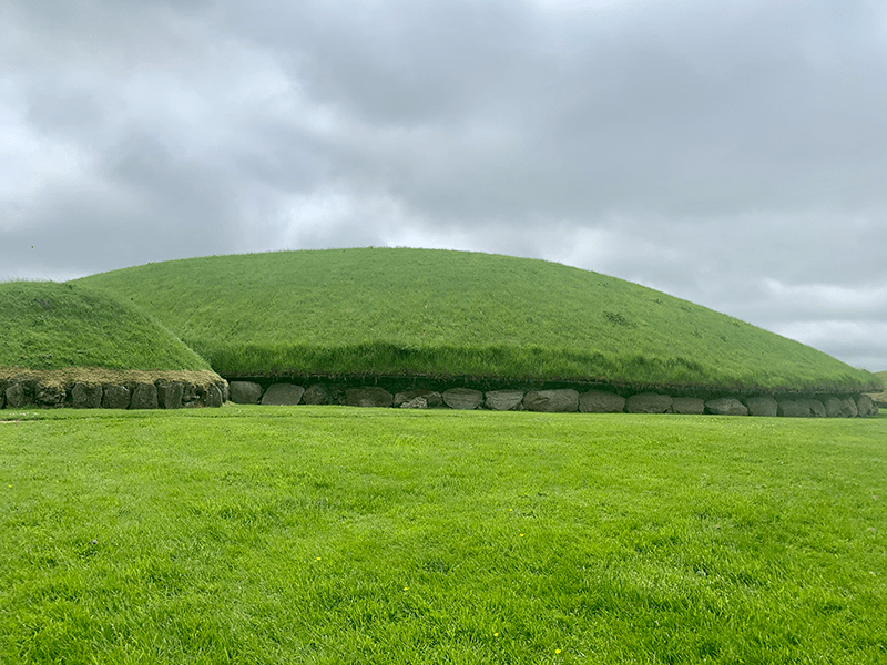 Two bright green sidhe mounds from the Knowth complex against a grey, cloudy sky.