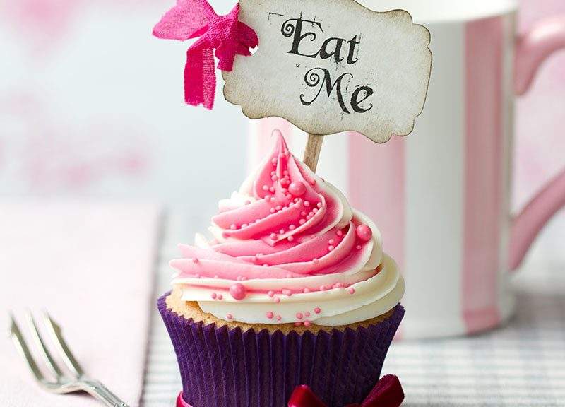 Beautifully pink and white frosted cupcake with a calligraphy sign with a bow that says "Eat Me" and a fork on a white and pink defocused background.