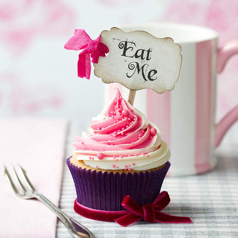 Beautifully pink and white frosted cupcake with a calligraphy sign with a bow that says "Eat Me" and a fork on a white and pink defocused background.