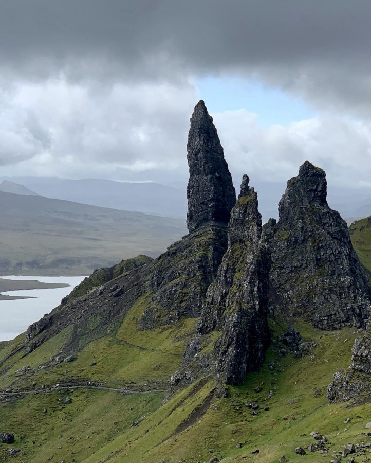 Old Man of Stór on the Isle of Skye in Scotland. Large, protruding shards of dark rock jutting out of a bright green hillside into a moody, cloudy grey sky with a hint of blue poking through the clouds