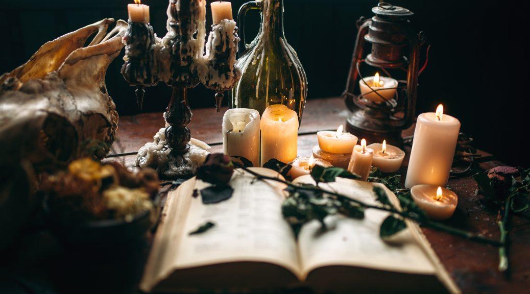 A spell book sits open with dried flowers laying across and several candles dripping on the table and in wine bottles with dramatic lighting.