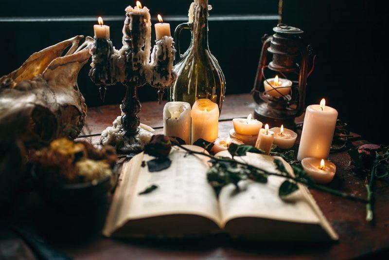 A spell book sits open with dried flowers laying across and several candles dripping on the table and in wine bottles with dramatic lighting.